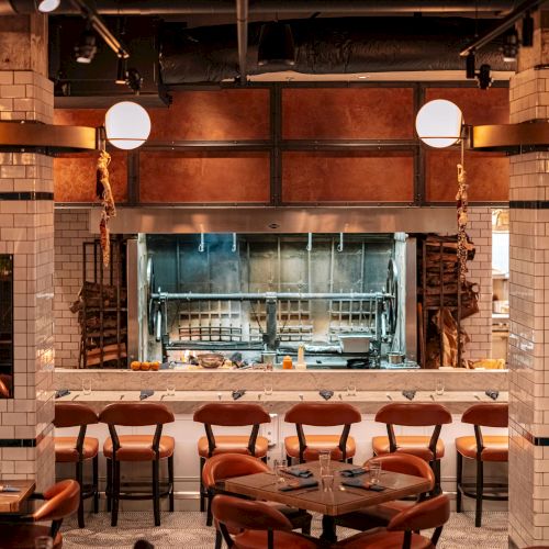 The image shows an upscale restaurant kitchen with open flame grills, elegant seating, and a chic industrial vibe with warm lighting and tiled walls.