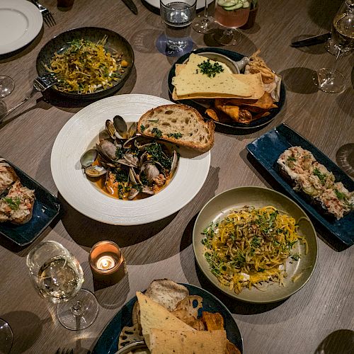 This image shows a table set for a meal with various dishes including pasta, bread, and seafood, along with plates, glasses, and a candle.