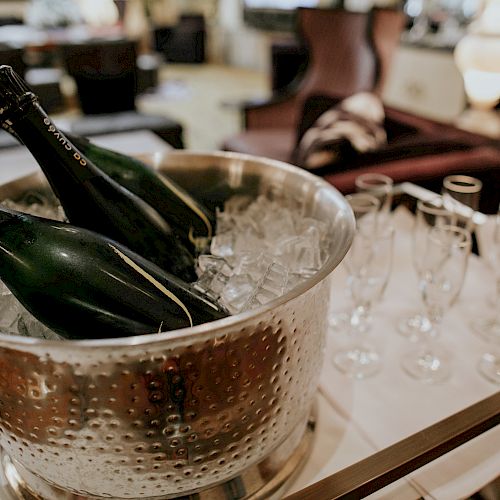 The image shows a silver bucket with ice holding three bottles of champagne, next to a row of empty champagne glasses, all set on a table.