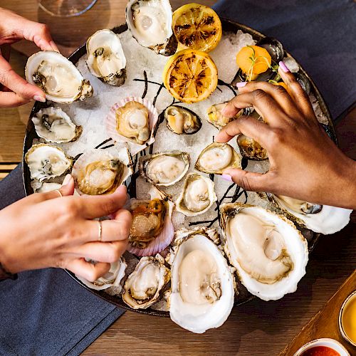 People are enjoying a platter of oysters with lemon and various sauces, as they reach in to pick oysters from the dish on a wooden table.