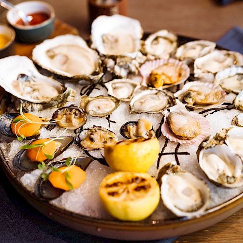 The image shows a large platter of assorted seafood, including oysters and clams, with lemon halves and garnishes on a bed of ice.