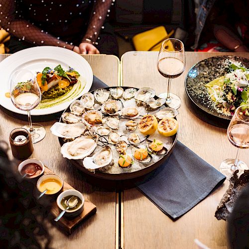 A restaurant table featuring seafood platter, dishes with pasta and salad, multiple sauces, wine glasses, napkins, and unseen people around it.