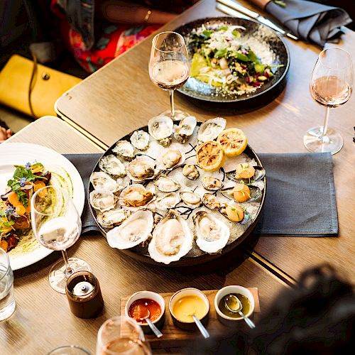 A group dining at a table with plates of oysters, salads, and wine glasses, accompanied by various sauces and a vibrant atmosphere.
