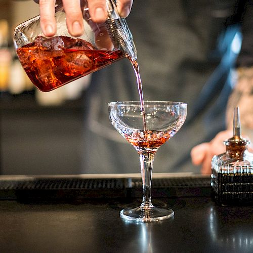 A bartender is pouring a red cocktail from a mixing glass into a coupe glass, with bottles and bar tools visible on the counter.