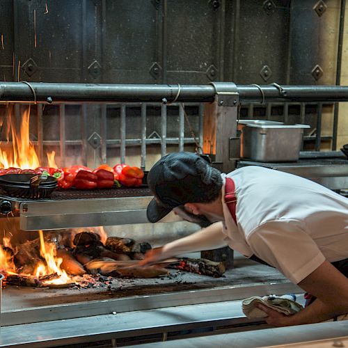 A person wearing a cap and white shirt is cooking over an open flame grill, adjusting food items like vegetables and possibly meat on the grill.