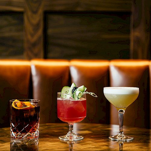Three cocktails are set on a wooden table against a background of brown leather seating.