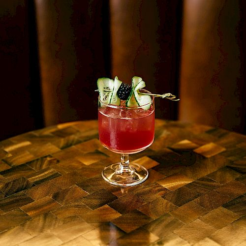 A glass with a red cocktail, garnished with cucumber slices and berries, is placed on a wooden table with a dark background.