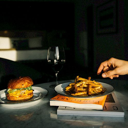 The image features a burger on a plate, a hand reaching for fries on another plate, a glass of wine, and two books on a table.