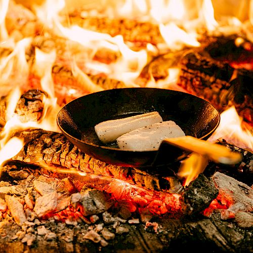 A frying pan with food is placed directly on a wood-burning fire, surrounded by flames and burning logs.