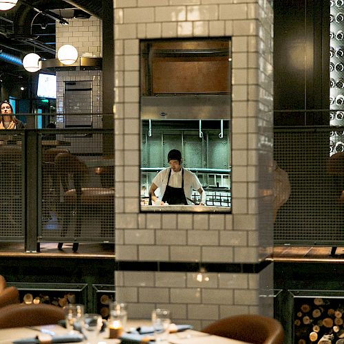 A restaurant kitchen view through a tiled wall with a chef working inside, in front of a seated dining area with tables and chairs neatly arranged.