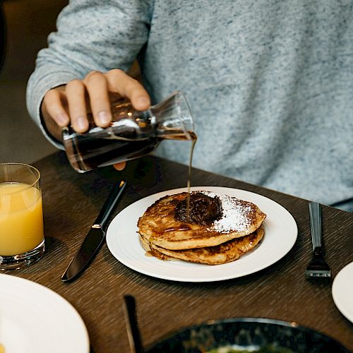 A person is pouring syrup over pancakes on a plate, accompanied by a glass of orange juice, with another dish also visible on the table.