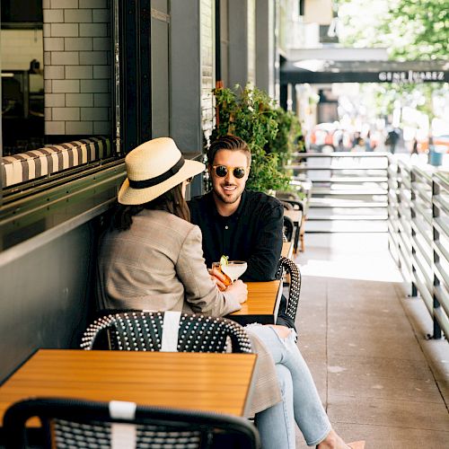Two people are sitting at an outdoor café, enjoying a conversation. One is wearing a hat, and the other is smiling and wearing sunglasses.