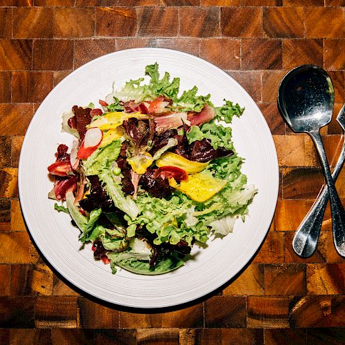 A fresh green salad served on a white plate, placed on a wooden table, with a fork and spoon beside it, arranged in a cross shape, ending the sentence.