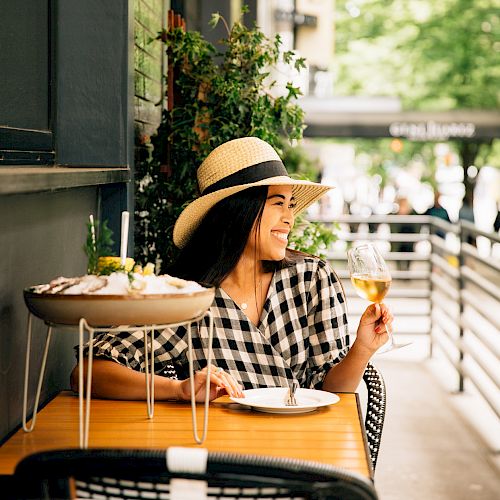 A woman in a hat and checkered dress sits at an outdoor table with food and a drink, smiling and enjoying the surroundings.