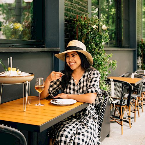 A woman in a checkered dress and hat sits at an outdoor café table, enjoying a meal with a drink, smiling, and surrounded by greenery.