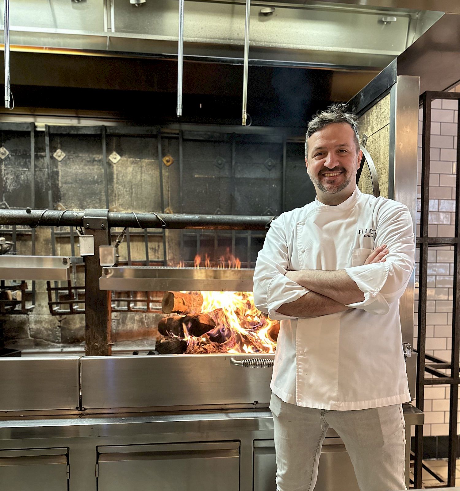 A chef in white uniform stands in a professional kitchen, arms crossed, smiling, with a flame visible behind.