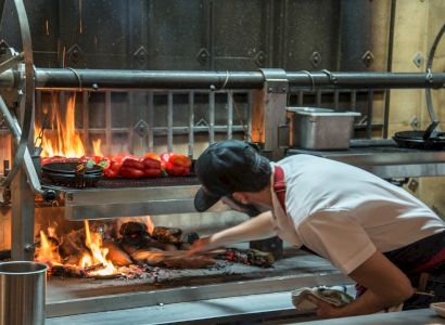 A chef, working in an industrial kitchen, tends to a fiery grill with vegetables on it, ensuring proper cooking.