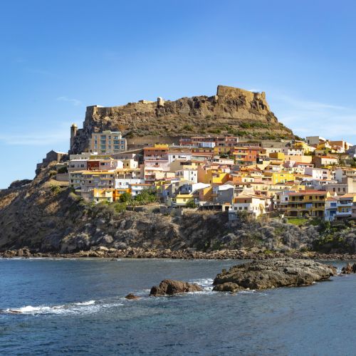The image shows a coastal town with colorful houses built on a rocky hill, crowned by an ancient fortress, bordered by a tranquil blue sea.